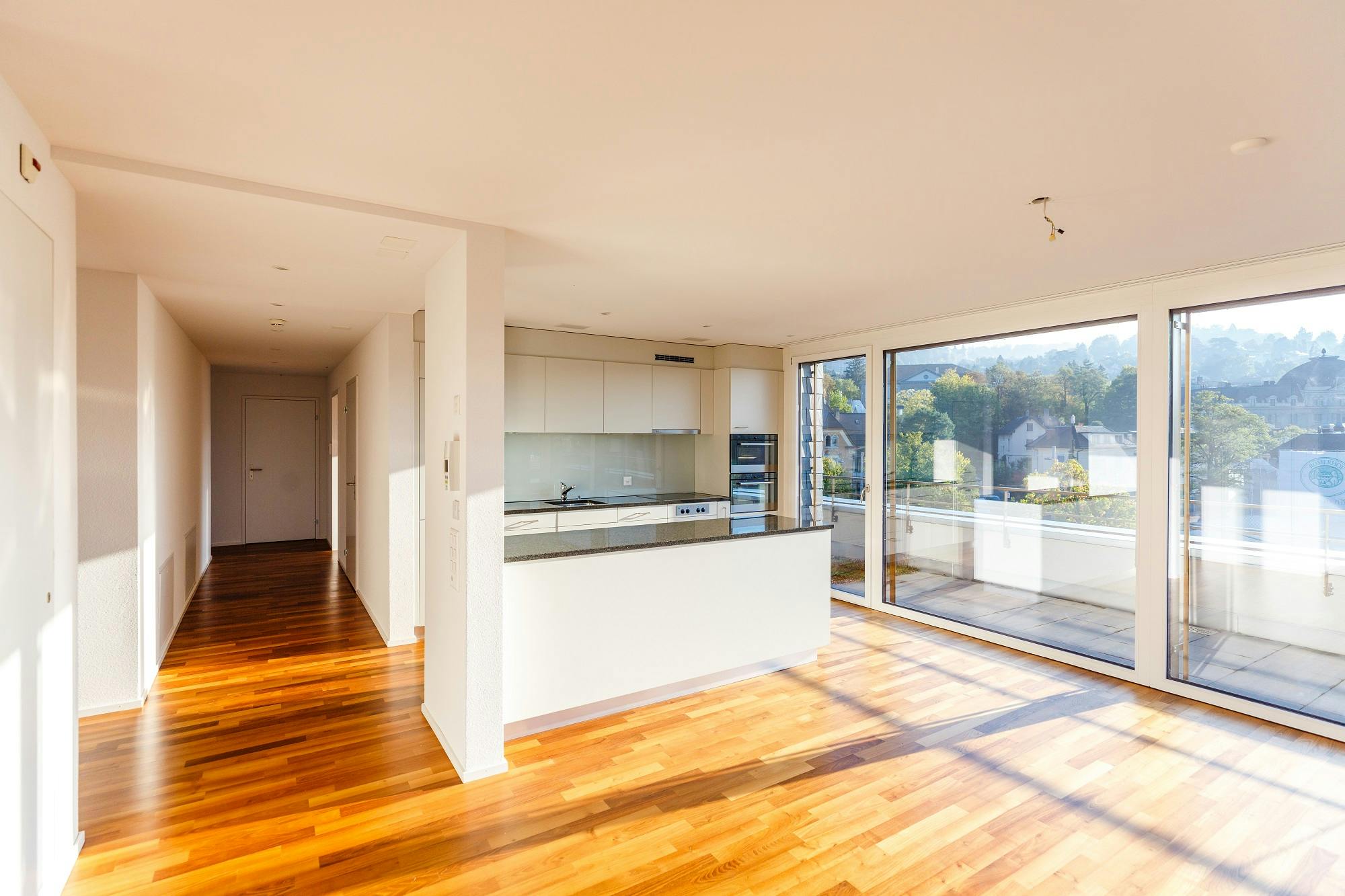Helle, moderne Küche mit Kücheninsel in einem offenen Wohnraum, Parkettboden, großen Fenstern und Balkonzugang.